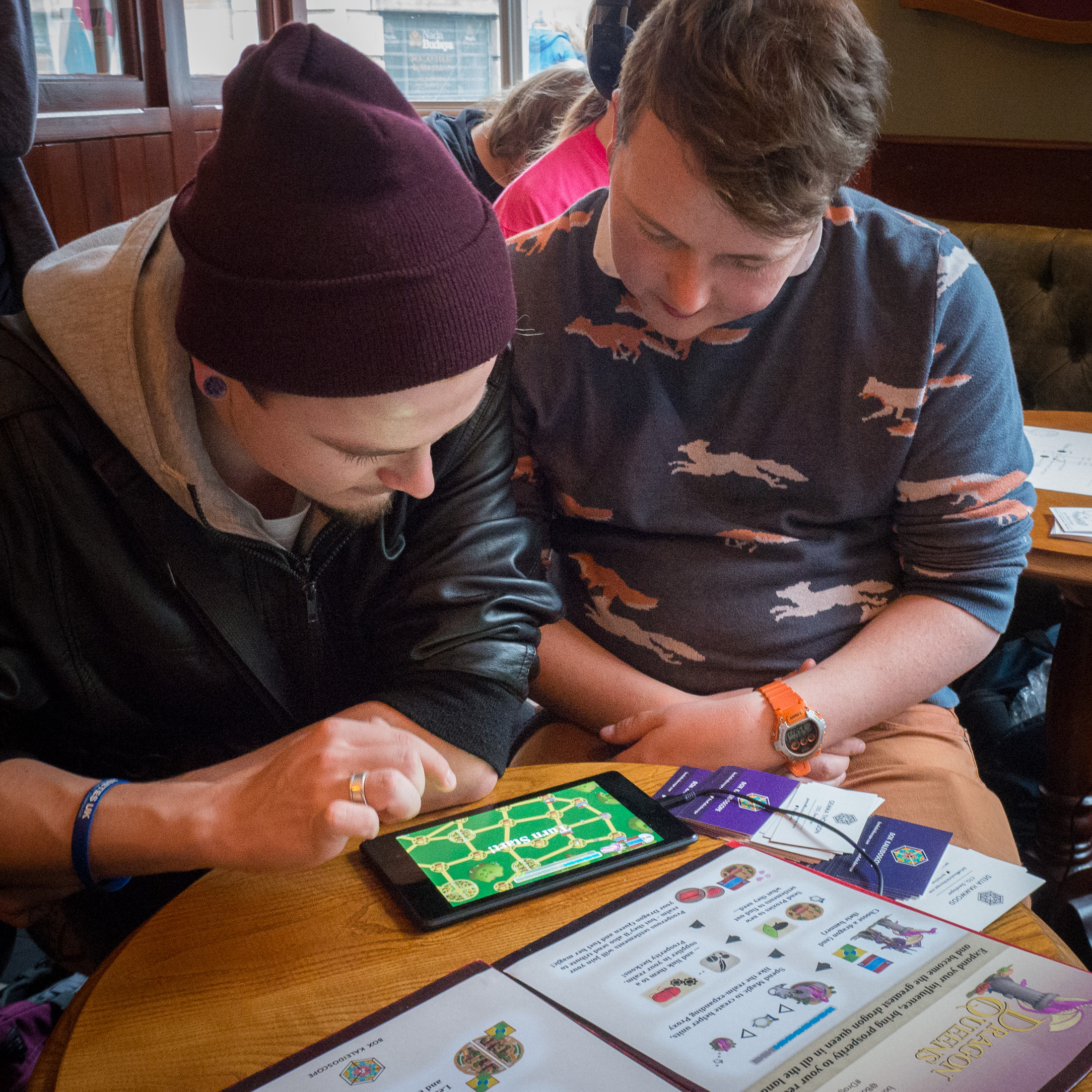 Two players at our booth, with a printed game guide visible on the table. One sits poised above the tablet, about to take their turn, while the other player observes.
